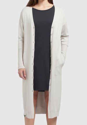 Mujer con un cárdigan largo abotonado color blanco roto sobre un vestido negro hasta la rodilla, con una mano en el bolsillo del cárdigan, sobre un fondo liso.