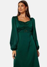 Zielona satynowa sukienka z kwadratowym dekoltem, marszczonym biustem, koronkowym wstawką i bufiastymi długimi rękawami, prezentująca gładką fakturę i elegancką sylwetkę.