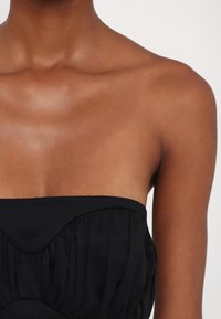 Rochie neagră fără bretele, cu o textură gathered, având un decolteu neted și un corset detaliat. Materialul pare ușor și fluid.