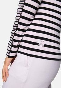 Detailaufnahme einer Person, die ein schwarz-weiß gestreiftes Oberteil mit Rüschendetails und eine helllila Hose mit sichtbarem Seitentasche trägt.