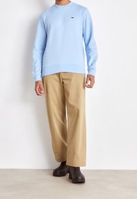 Felpa azzurra chiara con collo rotondo, polsini a costine e logo dorato. Abbinata a pantaloni beige a gamba larga e zoccoli marrone scuro.
