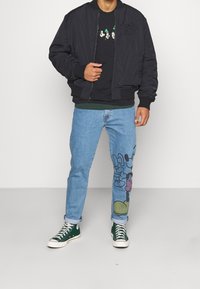 Svart bomberjacka med ribbade muddar, svart grafisk t-shirt, ljusblå jeans med en färgglad Mickey Mouse-print och gröna hög-top sneakers.