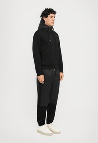 Svart zip-up hettegenser med en teksturert grå hette, kombinert med svarte bukser og hvite sneakers. Har sidelommer og en figursydd design.