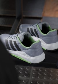 Paire de chaussures de sport grises avec des rayures noires et des touches vertes posée sur un banc sombre dans un vestiaire.