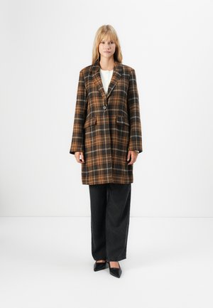 Blonde vrouw in een bruin en zwart geruit lang jasje, witte top, losse zwarte broek en zwarte puntige schoenen voor een witte achtergrond.