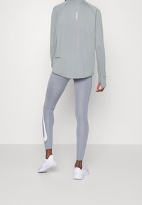 Ljusgrå långärmad sporttröja med hög krage och tumhål, kombinerad med grå leggings med vit logotyp och sportig design. Vita sneakers.