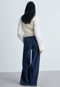 Canottiera a costine crema abbinata a una camicia bianca a maniche lunghe e a dei jeans scuri a gamba larga, con tasche posteriori e una vestibilità rilassata.
