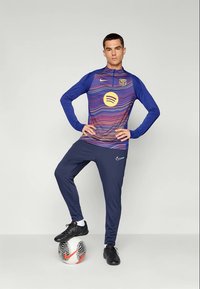 Nike sportska majica u plavoj boji s raznobojnim valovitom uzorkom, crne hlače i nogometne cipele, stoji na uzorkovanoj nogometnoj lopti.
