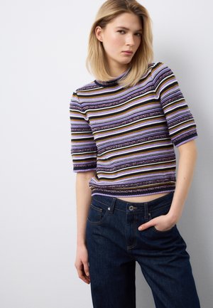 Giovane donna con capelli biondi indossa un maglione a maniche corte a righe viola, nere, bianche e gialle e jeans blu scuro, posando con la mano in tasca.