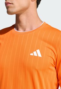 Πορτοκαλί αθλητικό πουκάμισο με υφασμένο, ριγωτό μοτίβο. Διαθέτει λευκό λογότυπο στο στήθος και στρογγυλή λαιμόκοψη με αντιθετική επένδυση.