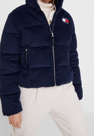 Osoba nosząca granatową sztruksową puchową kurtkę z logo Tommy Jeans, rozpiętą na białym golfie i jasnobeżowych spodniach.