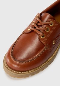 Brown leather shoe with a rounded toe, featuring gold eyelets and woven lacing. Textured upper with beige rubber sole. Stitch detailing visible.