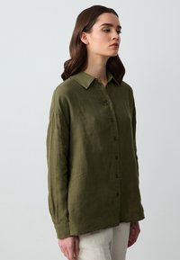 Camicia di lino verde oliva con colletto, maniche lunghe e chiusura a bottoni sul davanti. Presenta una vestibilità rilassata e una leggerezza sottile.