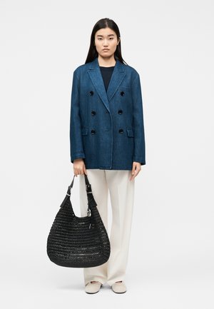 Femme portant une veste en denim bleu foncé à double boutonnage, un pantalon blanc à jambes larges, des chaussures blanches, tenant un grand sac à main noir tressé.