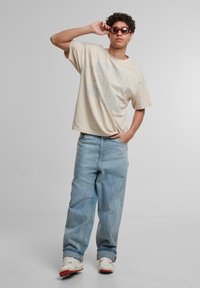 Beige oversized t-skjorte med trykt tekst, lyseblå baggy jeans og hvite joggesko med røde detaljer, iført solbriller.