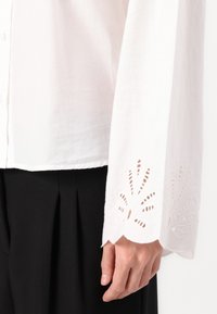 Hvit langermet skjorte med broderte utskjæringsdetaljer på mansjettene og knappestolpe, kombinert med svarte plysjbukser.