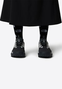 Svart och grå atletiska skor med texturerade sulor, som har grå sidor, runda skosnören och reflekterande detaljer. Svarta strumpor med logotyp.