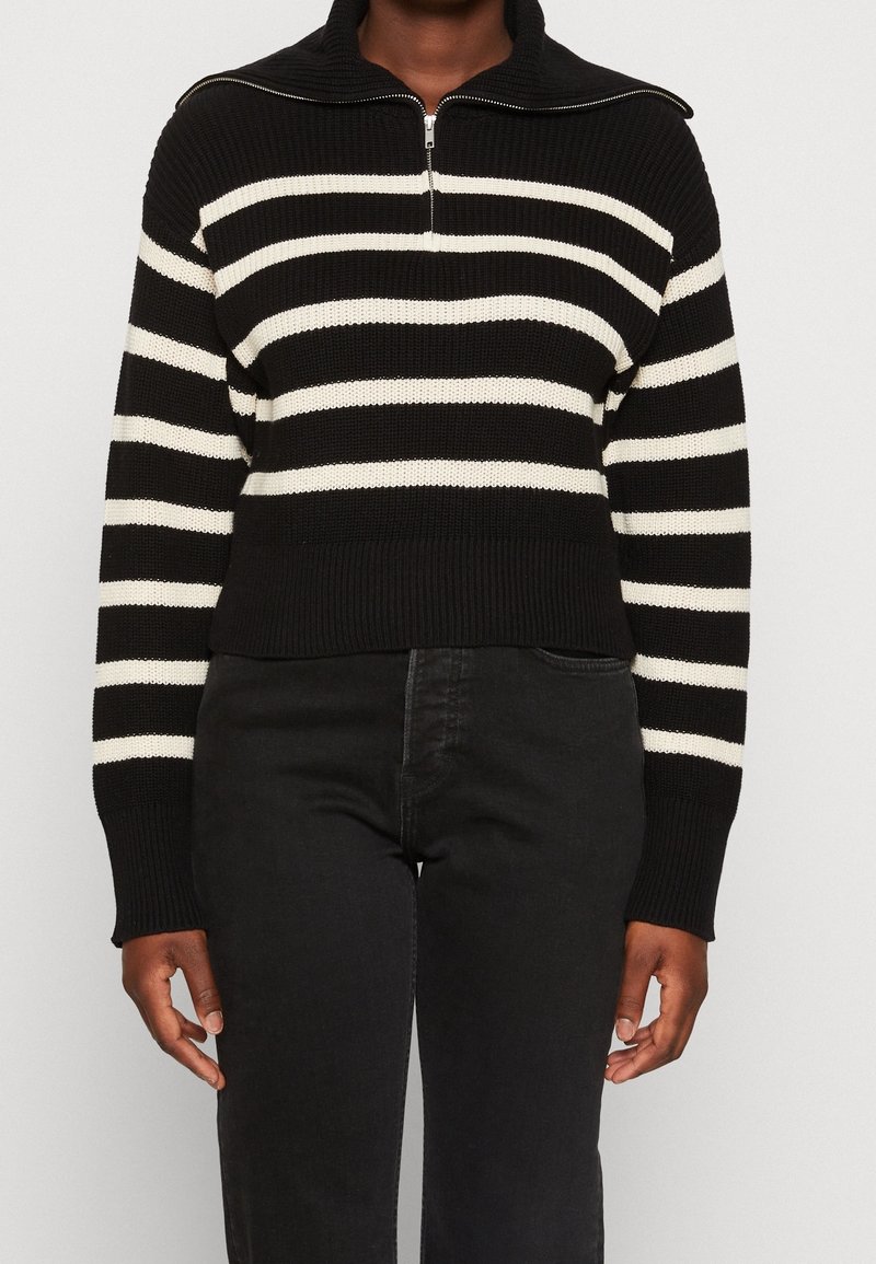 Personne portant un pull en maille rayé horizontal noir et blanc avec un col zippé et un pantalon noir taille haute.