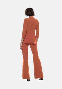 Blazer arancione su misura con pantaloni a palazzo coordinati, tessuto strutturato, maniche a tre quarti e colletto a punta. Calzature con tacco alto.