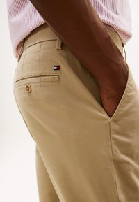 Nahaufnahme einer Person, die beige Hose mit einem kleinen rechteckigen roten, weißen und marineblauen Logo trägt und die Hand in der Vordertasche hat.