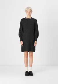 Robe pull noire avec un col rond et de longues manches ballons. Présente des poignets et un ourlet côtelés. Le modèle porte des chaussures noires et des chaussettes blanches.