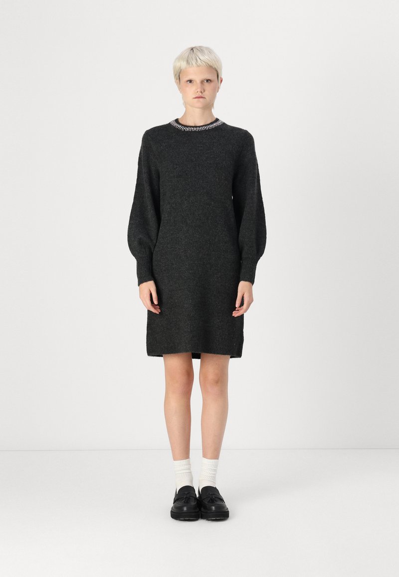 Robe pull noire avec un col rond et de longues manches ballons. Présente des poignets et un ourlet côtelés. Le modèle porte des chaussures noires et des chaussettes blanches.