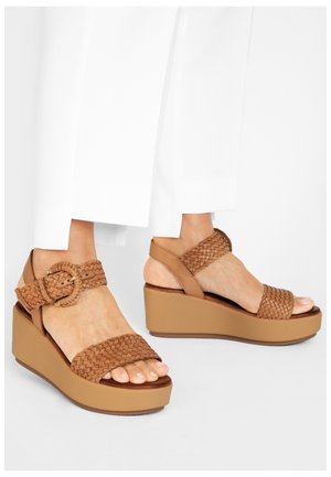 Pieds portant des sandales compensées tressées marron avec des brides et boucles à la cheville, assorties à un pantalon blanc raccourci sur un fond blanc.