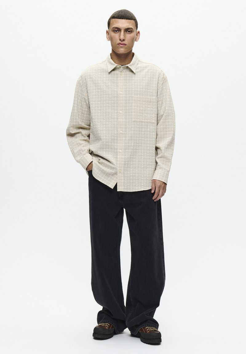 Chemise boutonnée beige à carreaux avec une poche, associée à un pantalon noir ample et des chaussures sombres avec des lacets marron.