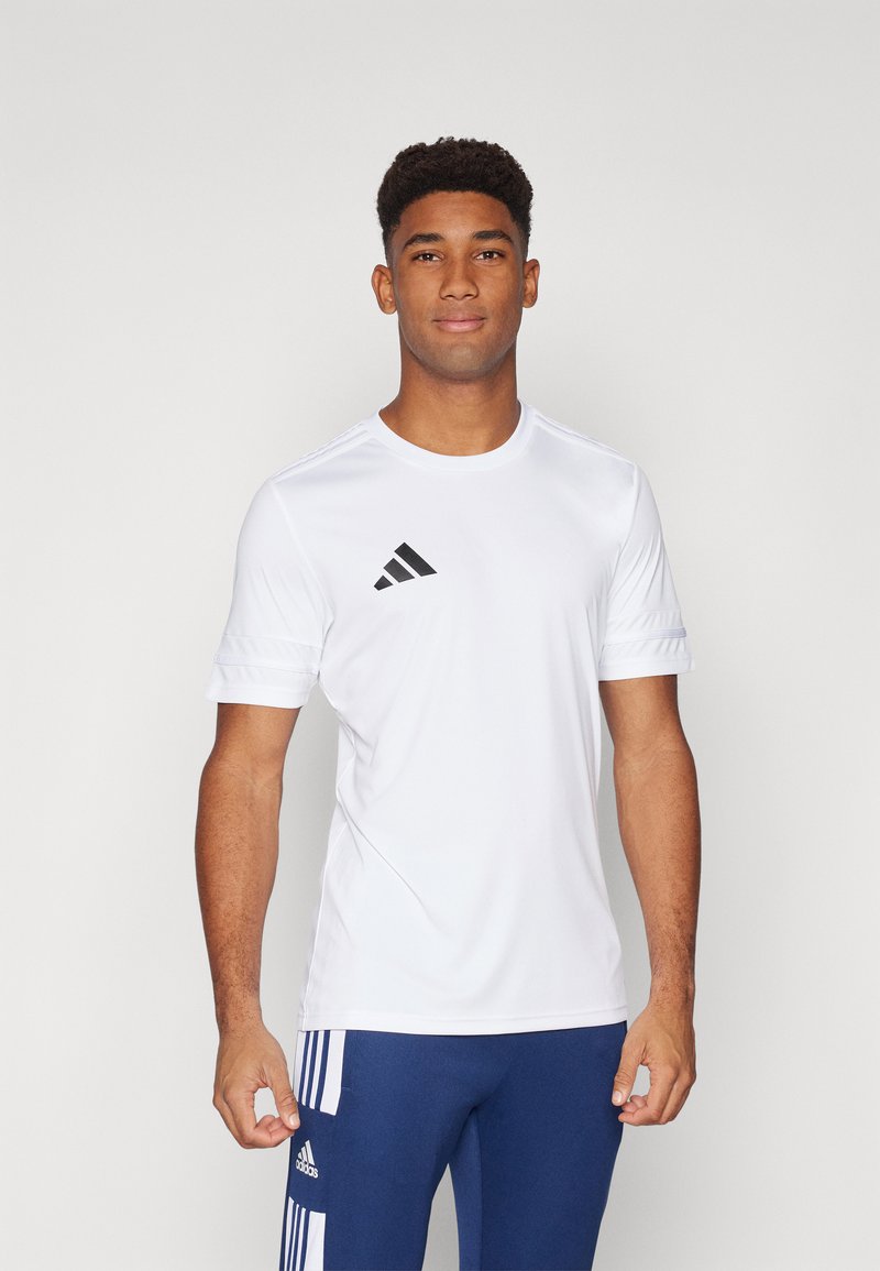 Camiseta deportiva de manga corta blanca con logo negro de Adidas. Tejido suave, cuello redondo y sutiles detalles en los hombros. Llevada con pantalones azules.