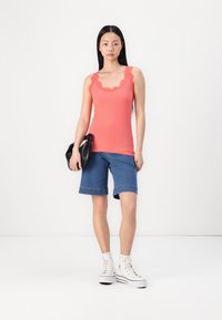 Top sin mangas de coral con cuello festoneado, combinado con unos pantalones cortos de mezclilla azul claro y zapatillas altas blancas. Bolsa negra en la mano.