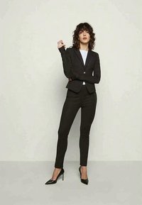 Melns pielāgots uzvalks, kas ietver piegulošu žaketi un šaurus bikses. Žakete ir ar iegrieztiem atloka jaciņām un vienu pogu, savienota ar melnām augstpapēžu kurpēm.