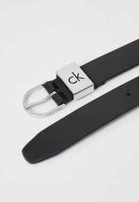 Ceinture en cuir noir avec une boucle métallique carrée gravée du logo "ck" et fermeture à double ardillon. Texture lisse avec des lignes épurées.