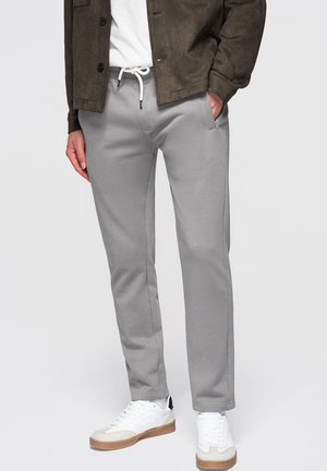 Person iført lysegrå snøre bukser, hvide sneakers, hvid skjorte og mørkebrun jakke med den ene hånd i lommen.