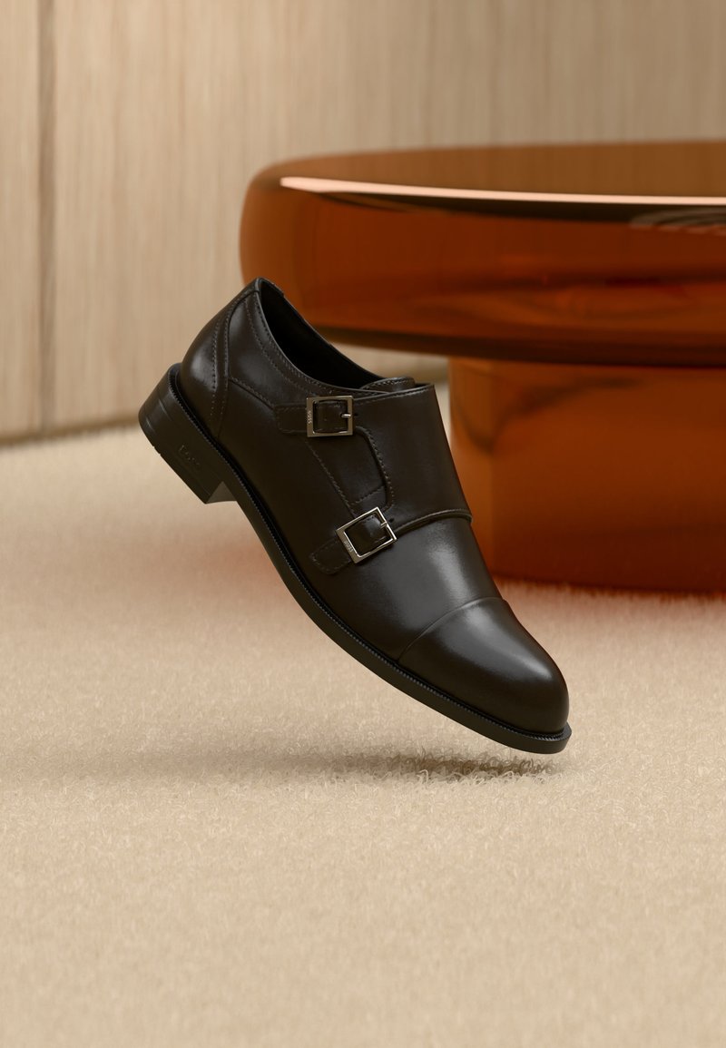 Chaussure noire en cuir à double boucle flottant au-dessus d'un tapis beige, avec du mobilier marron courbé en arrière-plan.