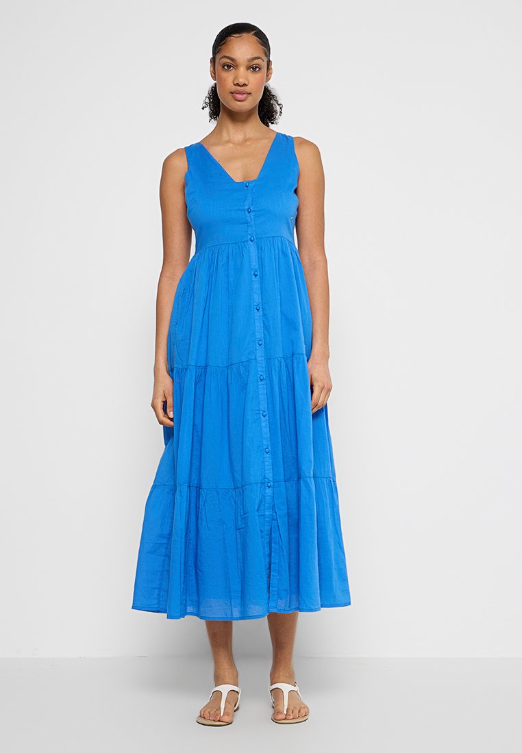 Mujer de pie con un vestido midi azul sin mangas y botones, y sandalias blancas en un fondo gris claro sencillo.