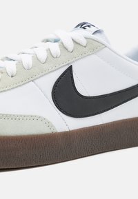 Zapatilla blanca de piel con acentos de ante gris claro. Presenta el logo Swoosh en negro, suela de goma texturizada marrón y cordones blancos.