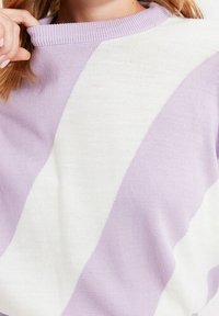 Detailní záběr na osobu v pleteném svetru s širokými diagonálními pruhy v pastelových fialových a bílých barvách, s vroubkovaným kulatým výstřihem, která drží pramen vlasů.