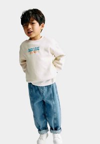 Sudadera color crema con texto gráfico azul, combinada con pantalones vaqueros azules holgados y zapatillas blancas. El conjunto presenta un ajuste relajado y puños enrollados.