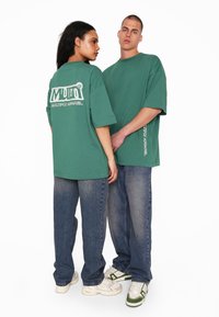 T-shirt verdi oversize con logo bianco stampato sulla schiena, abbinato a jeans in denim a vestibilità larga e sneaker, che mostrano uno stile streetwear casual.