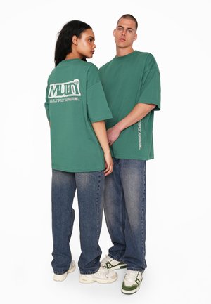 Zelené voľné tričká s bielym logom potlačeným na chrbte, kombinované s voľne strihanými džínsami a teniskami, ukazujúce ležérny streetwear štýl.