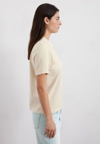Beige poloshirt met korte mouwen, klassieke kraag, gemaakt van katoen. Draagend met lichtblauwe jeans, bekeken vanaf de zijkant.