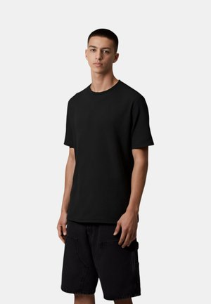 T-shirt nera a costine con collo rotondo, su uno sfondo chiaro, indossata con pantaloni corti neri dotati di tasche e dalla vestibilità rilassata.
