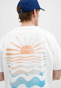 Witte katoenen T-shirt met een grote oranje zonafbeelding en blauwe golvende strepen op de achterkant, met een soepele combinatie van levendige kleuren.