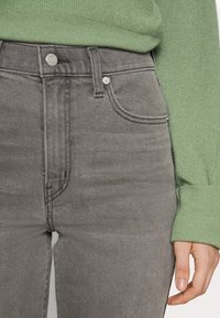 Jeans de mezclilla gris de cintura alta con una textura suave, diseño de cinco bolsillos y un suéter de canalé verde en contraste parcialmente visible.