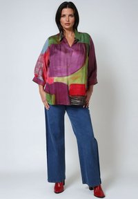 Satinbluse mit abstrakten Mustern in Lila, Grün und Rot, kombiniert mit weit geschnittenen blauen Jeans und glänzenden roten Knöchelstiefeln.