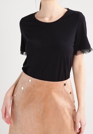 Mujer vistiendo una blusa negra de manga corta con detalles de encaje en las mangas y una falda de cuero color crema de talle alto con detalles de botones metálicos.