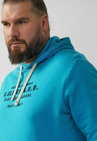 Turkusowy hoodie z białymi sznurkami, z nadrukowanym czarnym tekstem. Miękka tkanina o luźnym kroju, z kieszenią typu kangur.