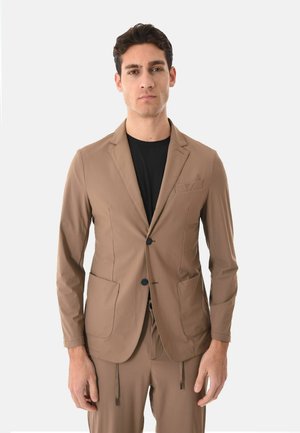 Uomo con blazer color beige e pantaloni abbinati, con camicia nera, in piedi davanti a uno sfondo chiaro e uniforme.