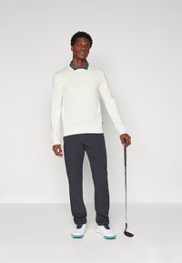 Maglione bianco sopra una camicia collare grigia, pantaloni scuri e scarpe da golf bianche con dettagli blu. Tiene un bastone da golf ed è in piedi su uno sfondo neutro.