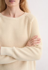Gebreid sweater van zachte, gebroken witte stof met een geribbeld textuur. Heeft een ronde halslijn en brede mouwen, met omgeslagen manchetten.